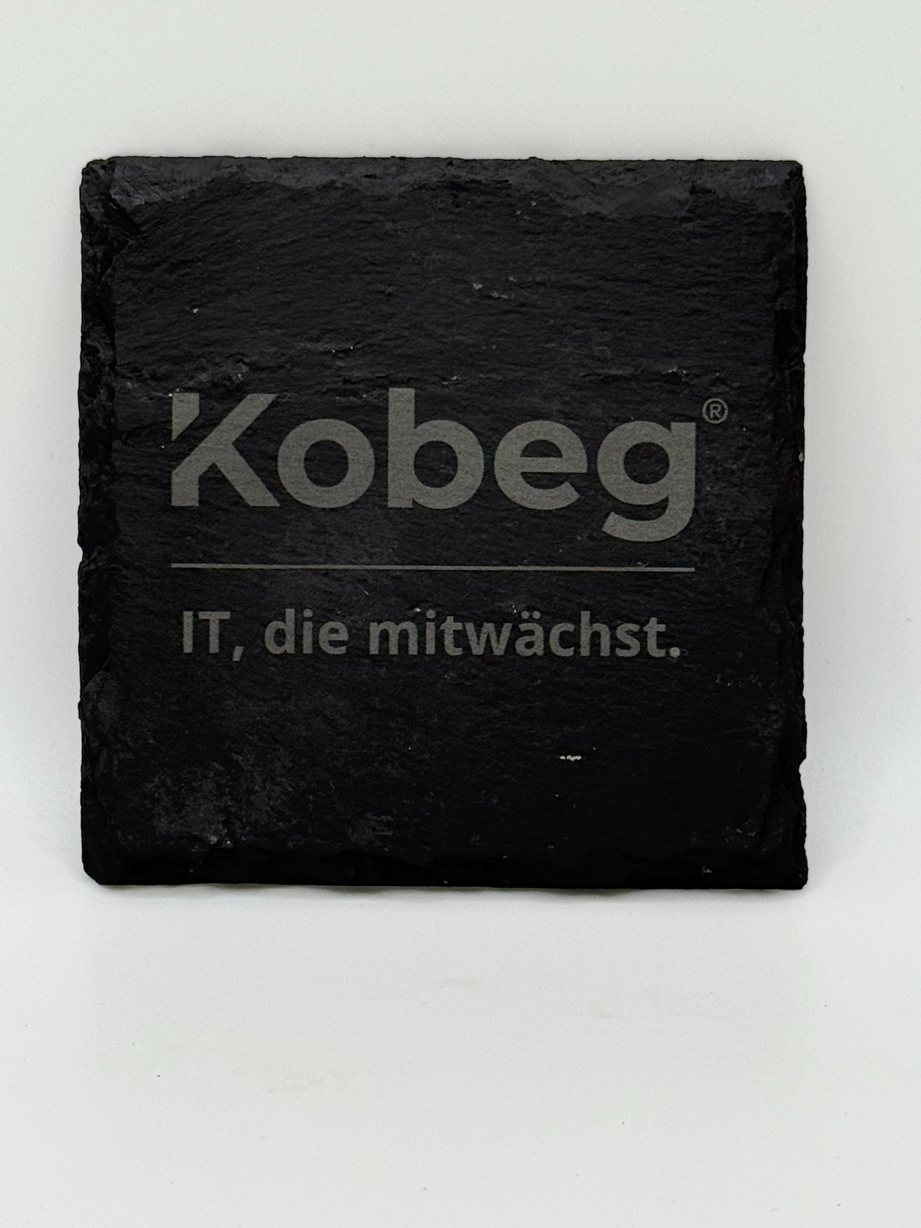 Eckiger Schieferuntersetzer mit lasergraviertem Firmenlogo Kobeg und Slogan "IT, die mitwaechst" als Beispiel fuer eine Logogravur